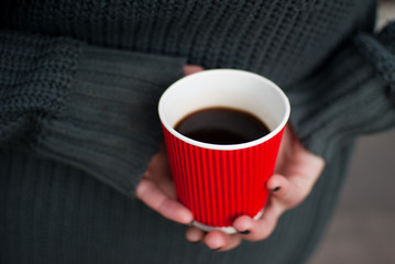 Girl wearing a gray sweater holding a red paper cup of black coffee in her hands. Cold spring days 