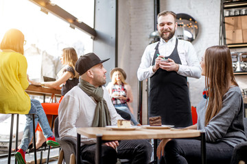 Waiter taking the order of a young couple sitting together in the beautiful cafe with other customers sitting on the background