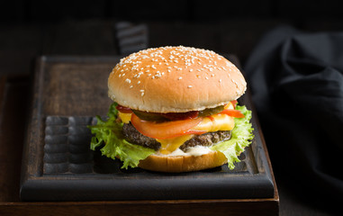 Delicious fresh tasty burger with beef, tomato, cheese and lettuce served on a wooden cutting board on dark background. Street fast food