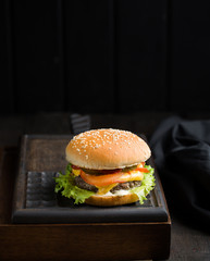Delicious fresh tasty burger with beef, tomato, cheese and lettuce served on a wooden cutting board on dark background. Street fast food