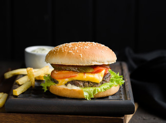 Delicious fresh tasty burger with beef, tomato, cheese, lettuce and french fries served on a wooden cutting board on dark background. Street fast food