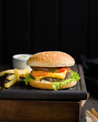 Delicious fresh tasty burger with beef, tomato, cheese and lettuce served and french fries on a wooden cutting board on dark background. Street fast food