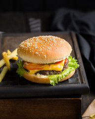 Delicious fresh tasty burger with beef, tomato, cheese, lettuce and french fries served on a wooden cutting board on dark background. Street fast food