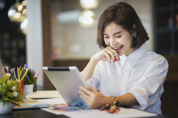 Young beautiful asian woman in white shirt holding pencil while looking at tablet