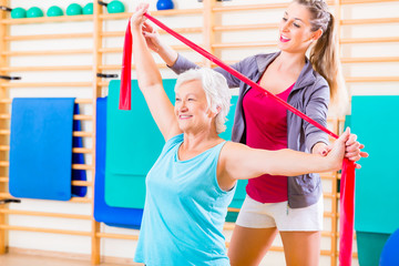 Senior woman with stretch band in fitness gym being coached by personal trainer