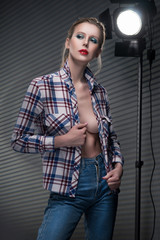 Beautiful slender blonde big breast girl without a bra wearing unbuttoned checkered shirt and jeans on a striped gray background near the studio lamp.