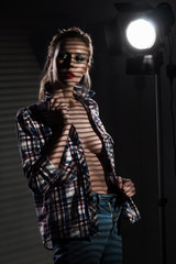 A beautiful slender blonde big breasts girl without a bra wearing an unbuttoned checkered shirt and jeans covered with shadows from the blinds near the studio lamp