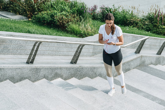 Summer day. Young woman athletes in white sportswear are standing on city street, steps, relax after sports training, use smartphone, listen to music. Girl checking email, browsing internet, chatting.