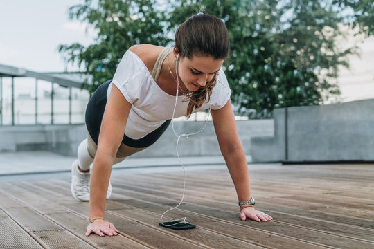 Summer day. Young woman doing sports exercises outdoors. Girl makes plank while looking at smartphone screen and listening to music. Workout, sports, sports training.