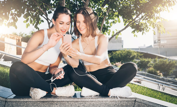 Two young women athletes in sportswear are sitting in park, relax after sports training, use smartphone. Girl shows her friend photos on smartphone screen. Online training, social network.