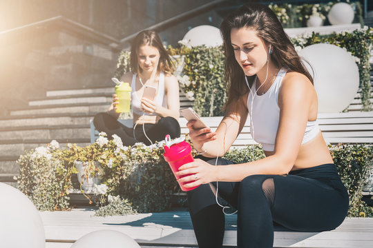 Sunny summer day. Two young women athletes in sports clothes are sitting on bench, relax after sports training, use smartphones, listen to music. Girls checking email, browsing internet, chatting.