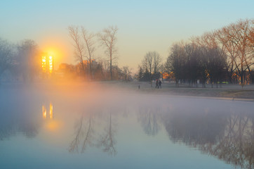 Two men in the early foggy morning walk their dogs. Male silhouettes in a thick morning fog on the river bank in the city park. Sunrays in the reflection of the windows of buildings cut through a mist