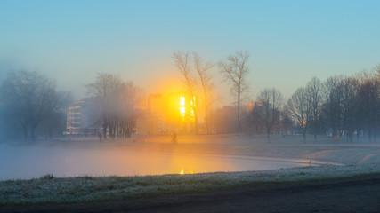 A lone silhouette of a cyclist riding in a strong fog along the river bank in a city park early in...
