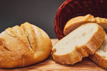 Fresh bread made from wheat flour in a wicker basket made of dark wood lies on a wooden board and against a dark background. Lighting from above through the hole. Slices of freshly baked bread.