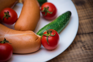 Cherry tomatoes, cucumbers and raw sausages on white plate on wooden table closeup