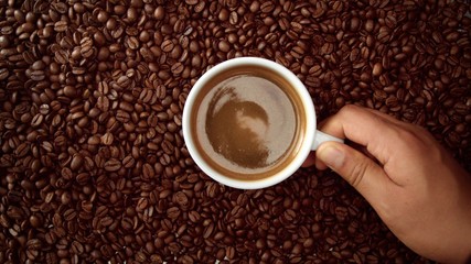 top view of hand with cup of black coffe put it on coffe beans background. White cup of black coffee