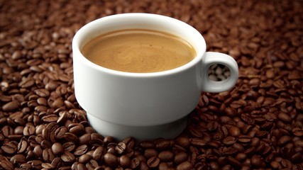 side view of white cup of black coffe on coffee beans. Coffee cream moving in cup