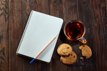 Cup of tea with cookies, workbook and a pencil on a wooden background, top view