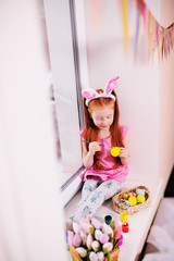 a small pretty girl with red hair with rabbit ears on her head is painting eggs for an Easter basket on the windowsill against the window