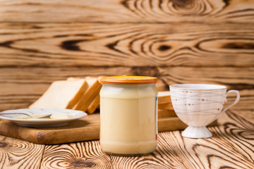 A bowl and a bottle of condensed milk and a teaspoon on a wooden table with background of toasts and tea cup