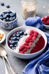 Smoothie bowl with fresh raspberry, blueberry, coconut flakes and chia seeds. Grey stone background.