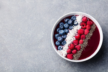 Smoothie bowl with fresh raspberry, blueberry, coconut flakes and chia seeds. Grey stone background. Top view. Copy space.