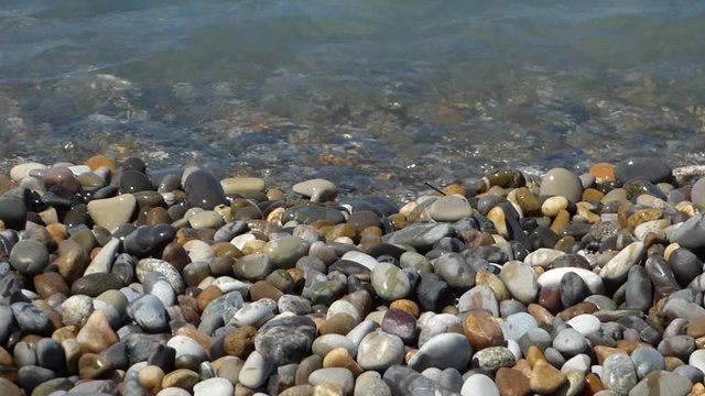 Sea surf on multicolored polished stones and pebbles. Water waves on beach, closeup view. Sunny summer day. Natural background.