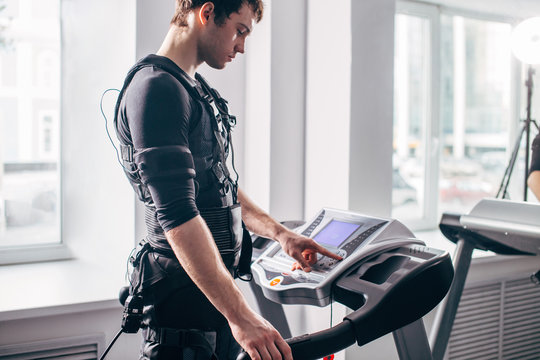 ems training. Fit Man wearing black electromiostimulation suit preparing for running on treadmill near window