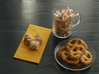 Homemade cookies on a transparent glass plate and marshmallows on napkin and in the cup on a wooden table