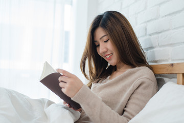 Lifestyle happy young Asian woman enjoying lying on the bed reading book pleasure in casual...