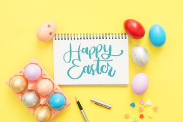 Flat lay top view colorful easter egg painted in pastel colors composition and text Happy Easter Day on notebook with paint brush on yellow pastel color background. Top view Easter day background.
