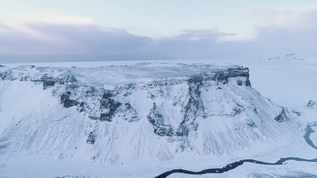 Aerial shot flies straight over a snow covered mountain with a creek winding alongside. Pristine winter environement at sunrise. Ocean in the far distance.
