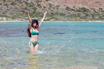 Portrait of a cheerful young girl in a black hat and green swimsuit standing in the emerald sea water and playing with water, making splashes, copy space