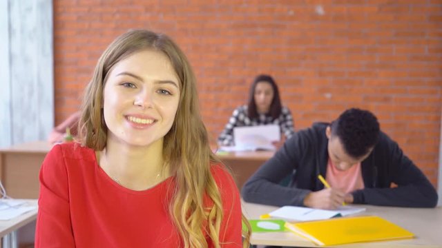 young attractive blond woman sitting opposite computer and smiling happily at camera. good-humored face of a student. In the background, young people work 4k