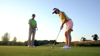 Low-angle side view of an attractive fit woman exercising hitting technique during golf class with an experienced professional player in summer - Powered by Adobe