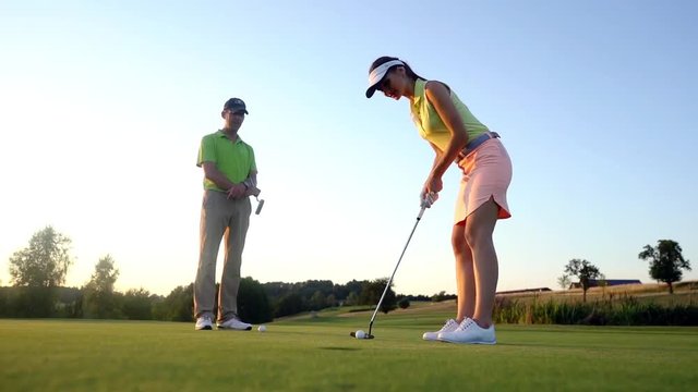 Low-angle side view of an attractive fit woman exercising hitting technique during golf class with an experienced professional player in summer