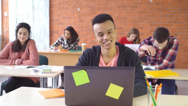 young attractive man sitting opposite computer and smiling happily at camera. good-humored face of a student. In the background, young people work 4k