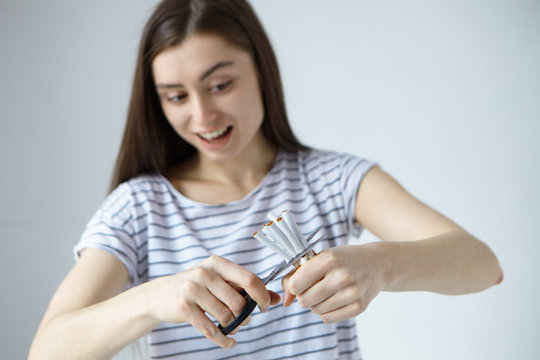 Horizontal shot of cheerful young woman dressed casually holding scissors and cigarettes, cutting them in halves, having happy expression of winner who could break bad habit. Stop smoking concept