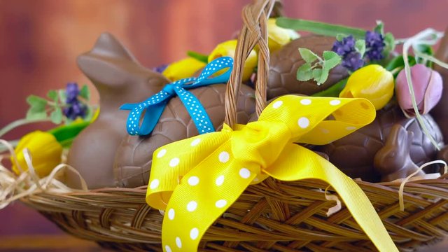 Happy Easter hamper of chocolate eggs and bunny rabbits in large basket with silk tulips on dark wood table, panning macro.