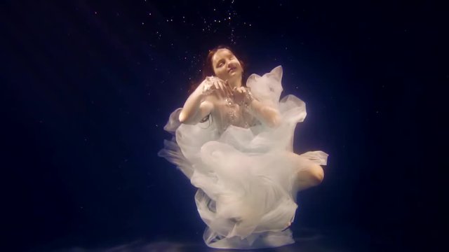graceful little girl with red hair and in white dress ia moving under water, embodying image of princess
