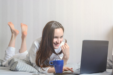 A young woman in pajamas is lying in bed and chatting on social networks through a laptop. Modern youth