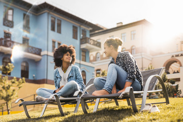 Black girl with curly Afro hair is having a chat with her female smiling Caucasian friend while both sitting on deck chairs on the green fresh lawn between modern houses on a spring sunny day - Powered by Adobe