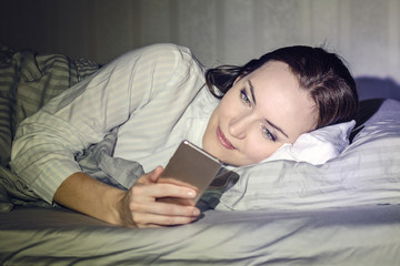 The girl lies on the bed on her side and looks at the smartphone. Shot from the side in a dark key