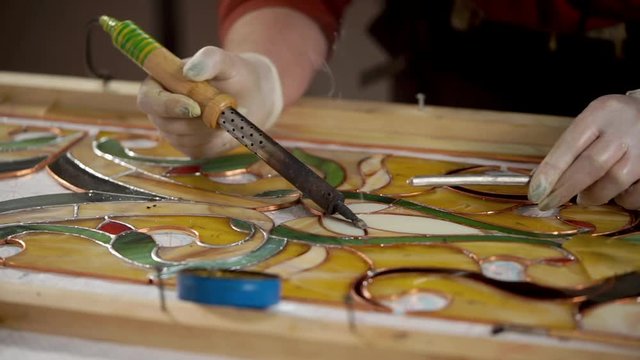 Master is using a soldering iron for decorating and processing frame of stained-glass picture. He is melting tin and applying it on copper base.