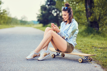 Happy young woman sitting on longboard and using phone outdoors