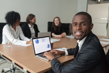 Smiling african-american businessman in suit looking at camera at diverse team meeting, business analyst using laptop with project statistics at screen, professional black manager at work portrait