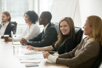 Young smiling businesswoman looking at camera at corporate group meeting, business training participant sitting at conference table with diverse colleagues, company manager portrait at team briefing