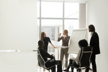 Diverse business team listening to female speaker discussing presentation on flip chart at meeting...