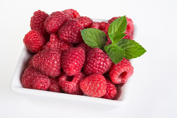 Red raspberries with mint on white background. Beautiful close-up of juicy sweet berries in a square bowl with decorative green peppermint leaves on the table.