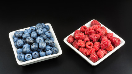 Healthy juicy raspberries and blueberries. Decorative red and blue forest berries in white square bowls on a black background.